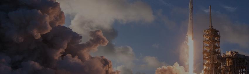 A rocket lifts off from its launch pad with clouds of smoke billowing around it, set against a blue sky.