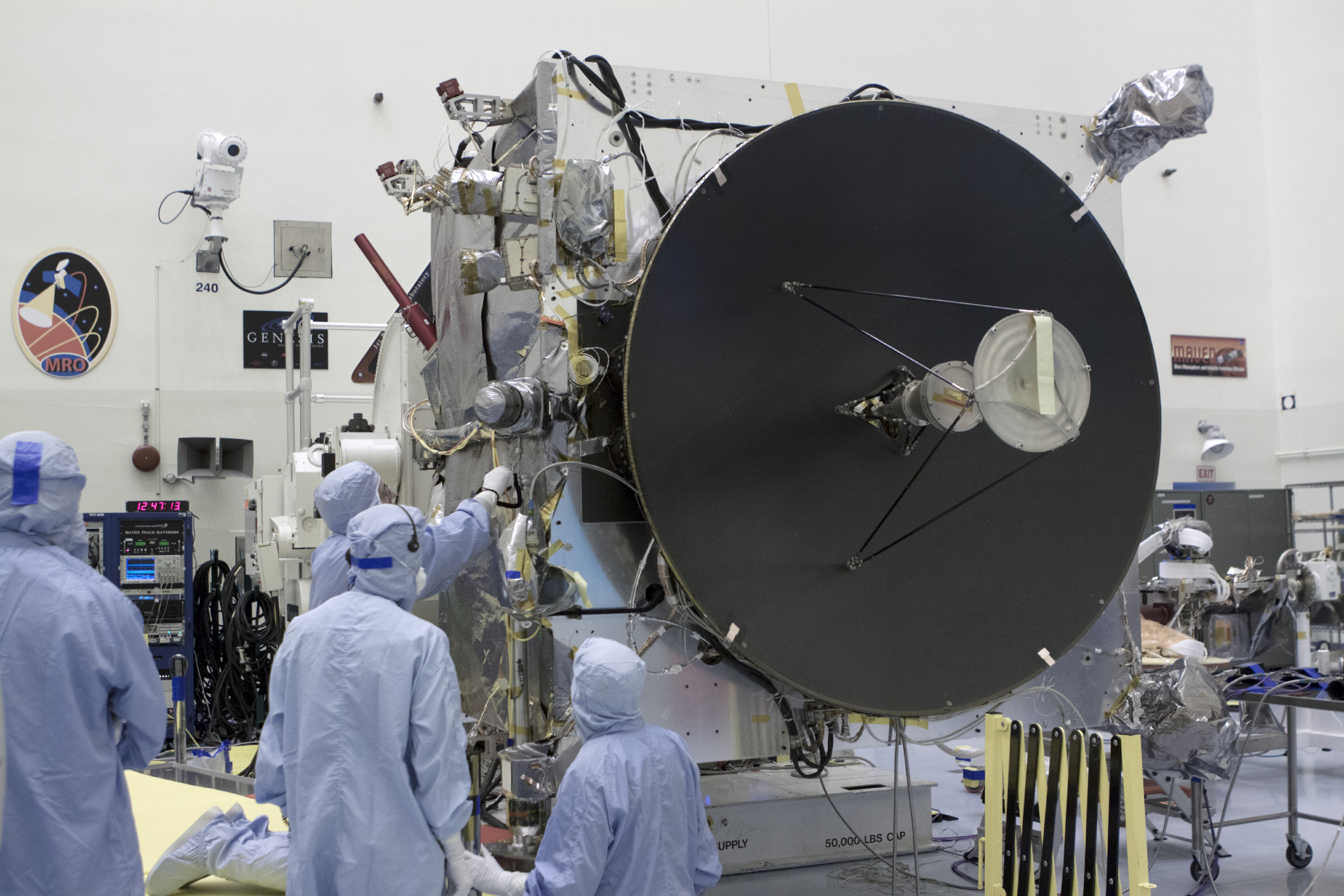 Technicians in cleanroom suits work on a spacecraft with a large dish-shaped antenna in a laboratory setting