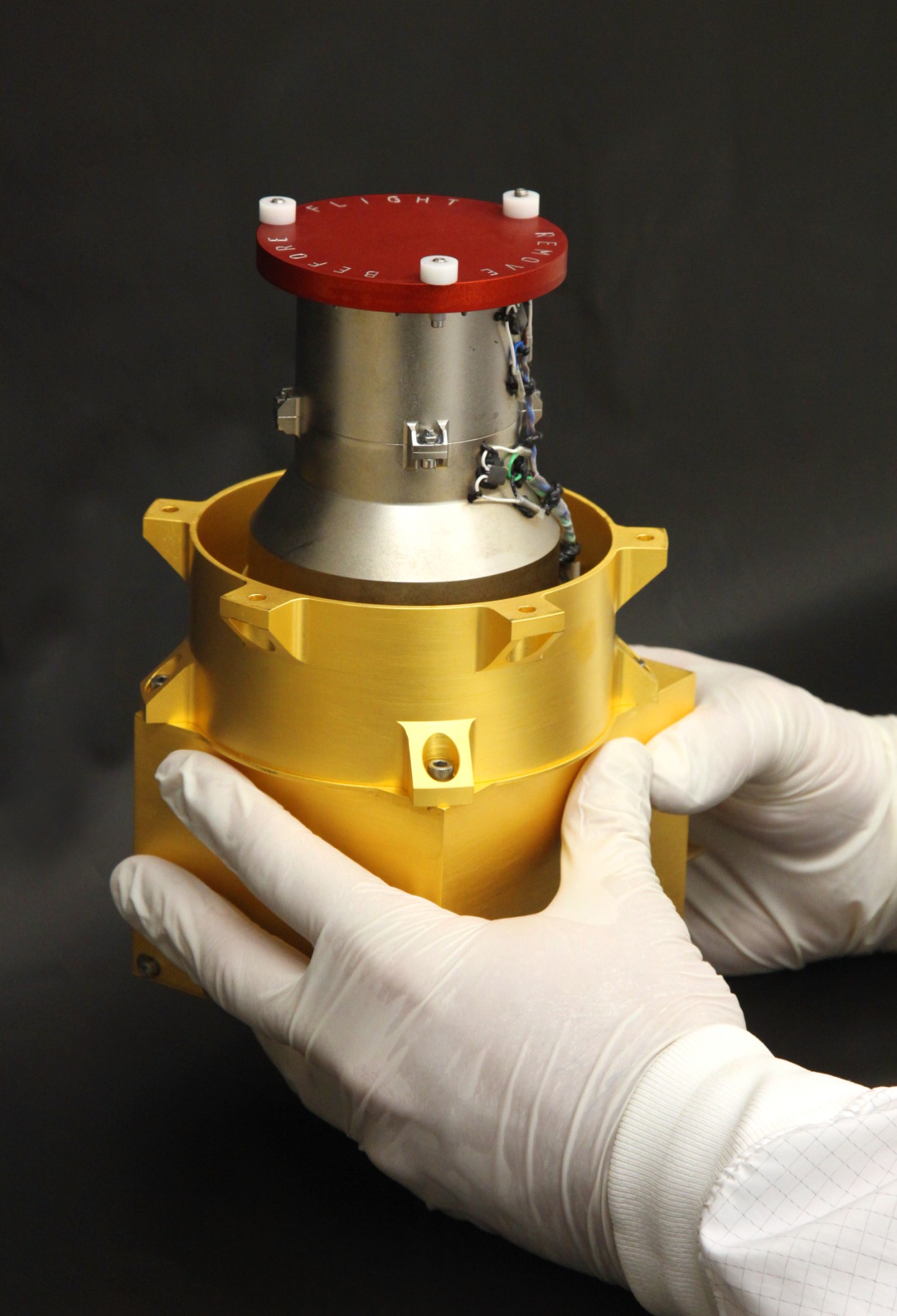 A person wearing white gloves holds a metallic device with a red cap and yellow casing