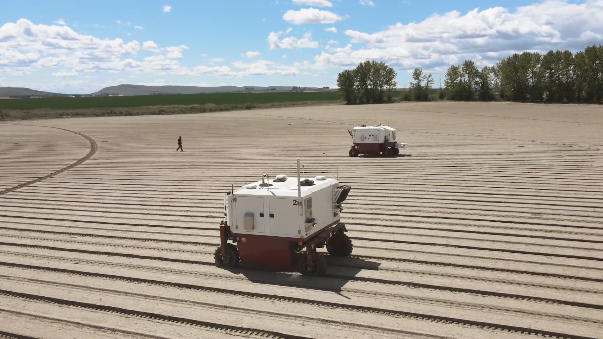 Two autonomous agricultural vehicles traverse and work on neatly plowed fields under a partly cloudy sky, while one person is seen walking in the background. Trees and distant hills border the field.