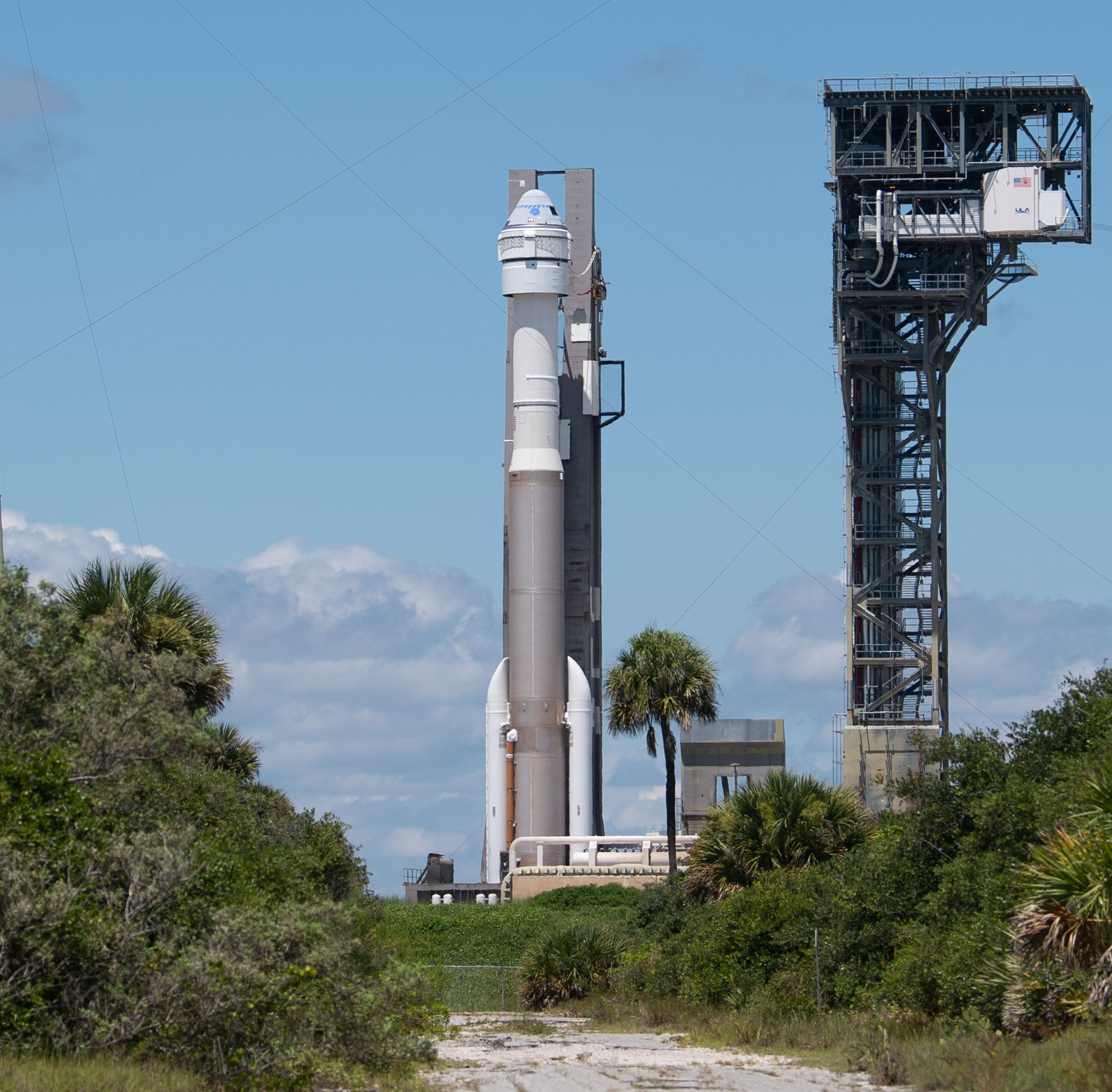 A space rocket stands vertically on the launch pad, surrounded by greenery and a blue sky with clouds, next to a tall support structure.