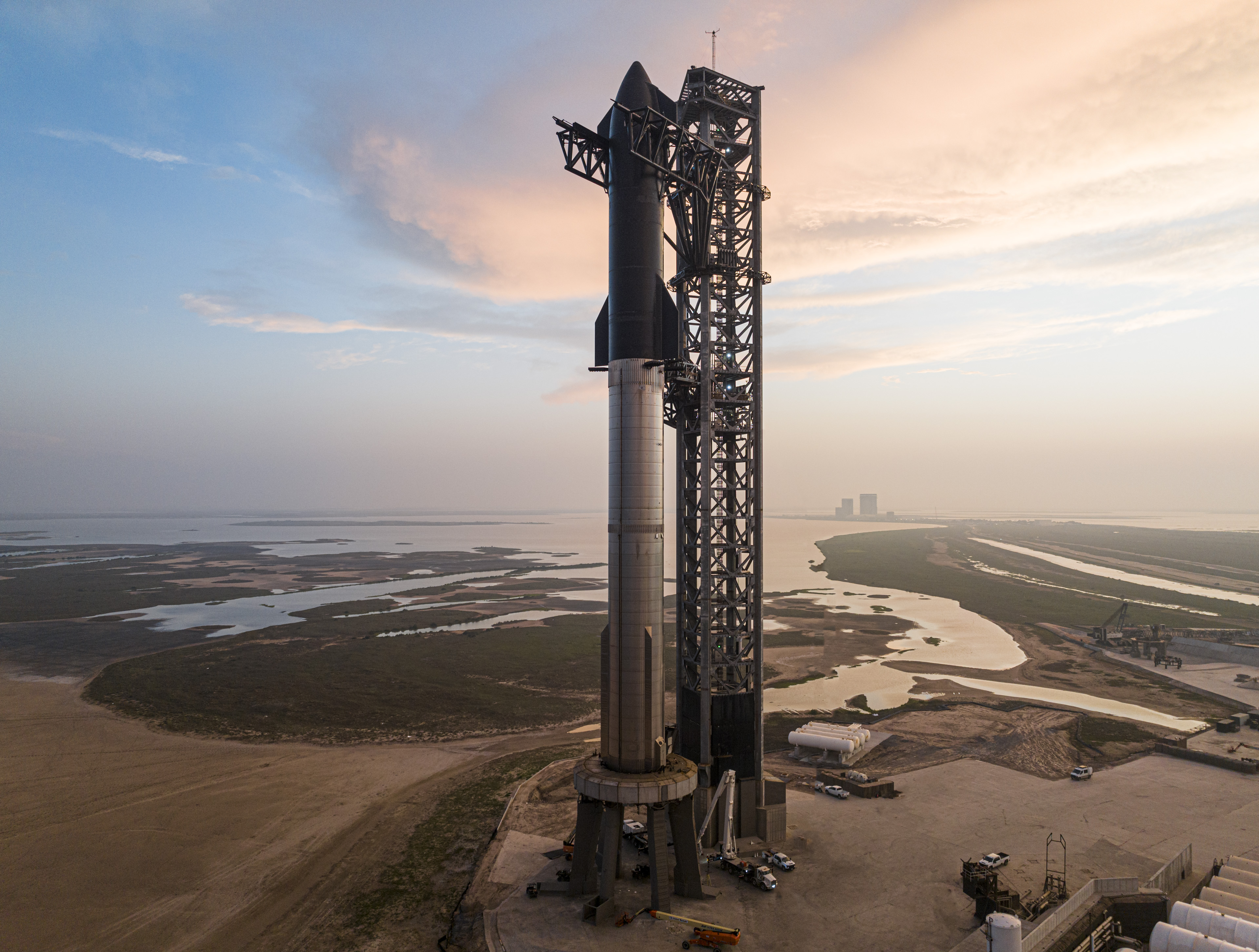 A large rocket stands vertically on a launch pad with support structures nearby. The surrounding area includes water and flat, open land extending into the distance under a twilight sky.