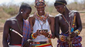 Three people in traditional attire look at a smartphone together outdoors, holding long sticks decorated with colorful patterns.