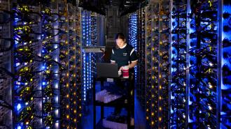 In a bustling data center, a person works with a laptop on a cart, surrounded by racks of illuminated servers and cables.