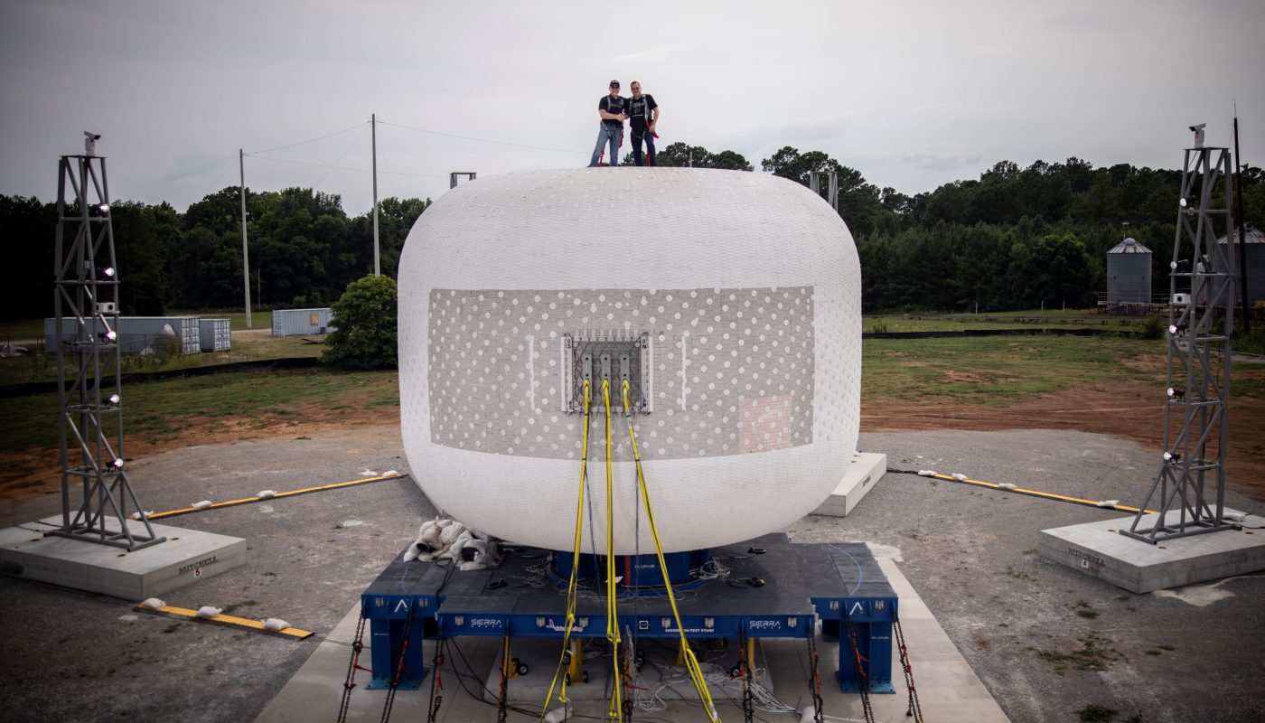 Two people stand atop a large, white, dome-shaped structure with cables attached to its sides. The structure is situated outdoors, surrounded by greenery and various equipment.