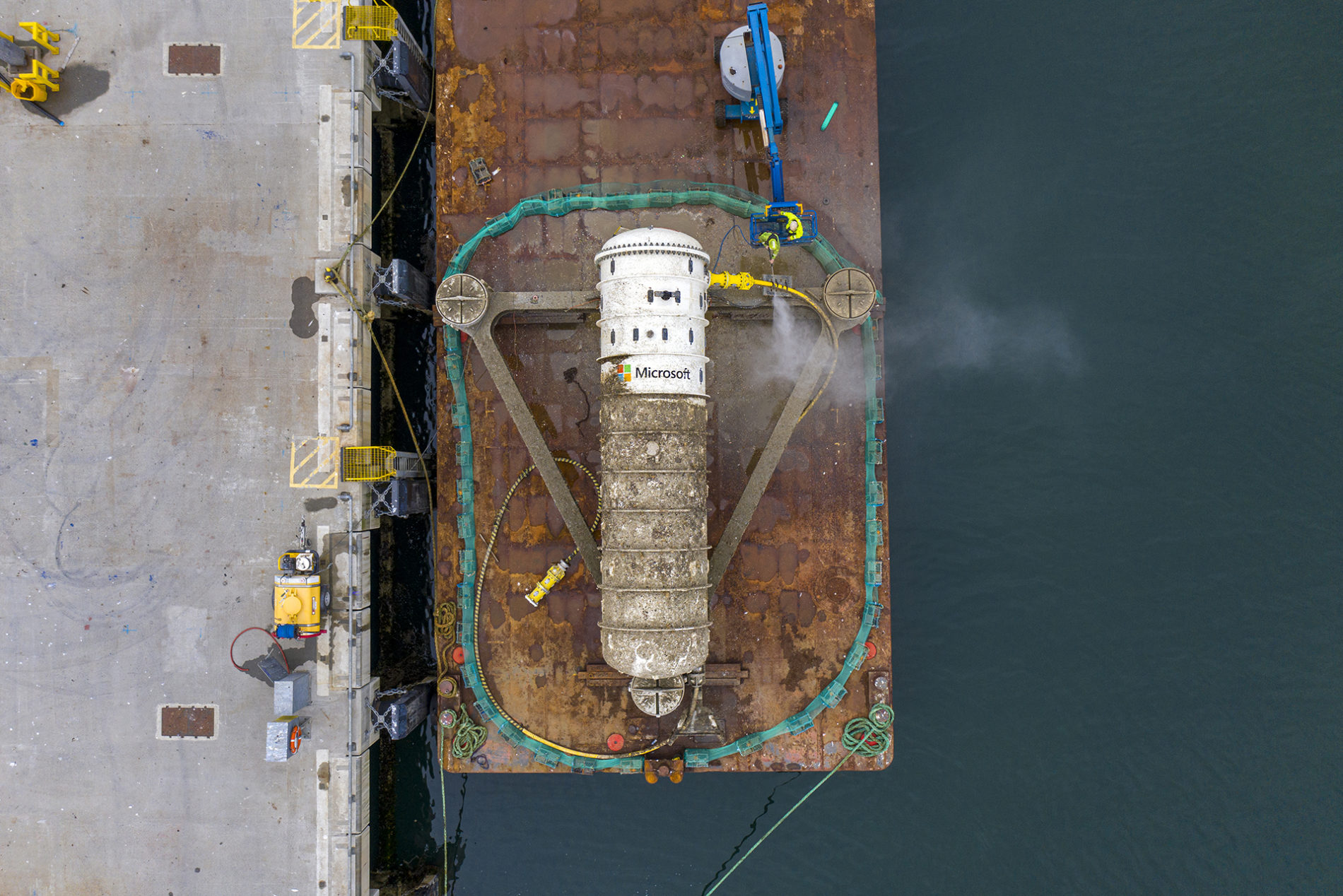 Aerial view of a rusted industrial cylinder labeled "Microsoft" on a barge at a dock, with water and dock equipment visible.