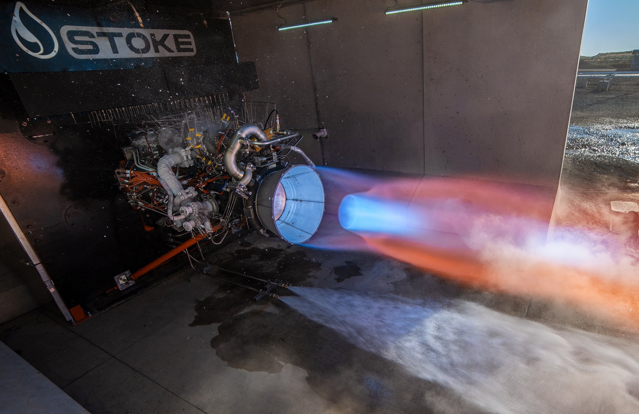 A rocket engine undergoing a test, emitting a bright blue and orange exhaust flame, in a controlled environment with the word "STOKE" visible on machinery.