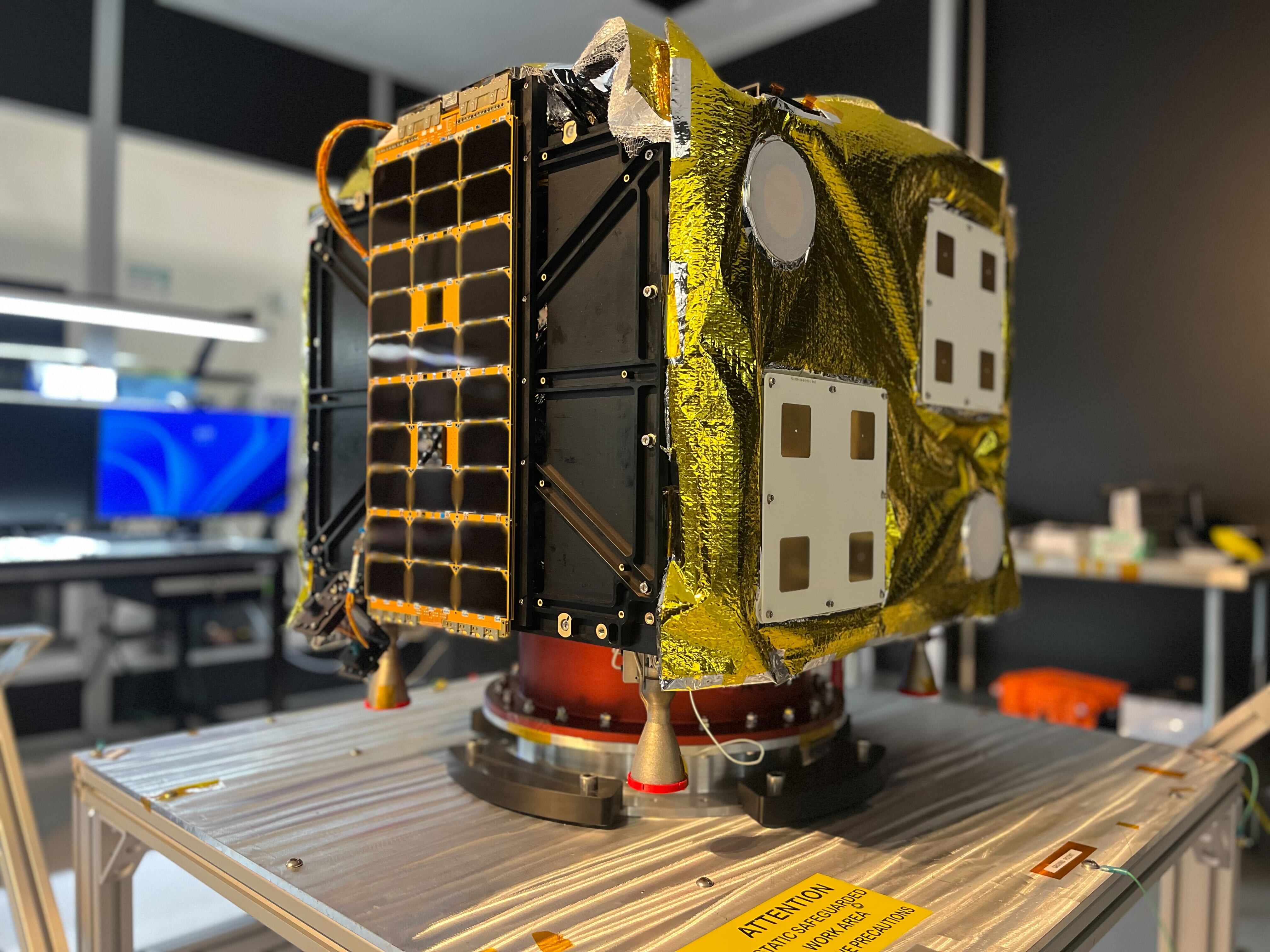 A small satellite with gold foil insulation and various sensors mounted on its surface is displayed on a metallic platform in a laboratory setting.