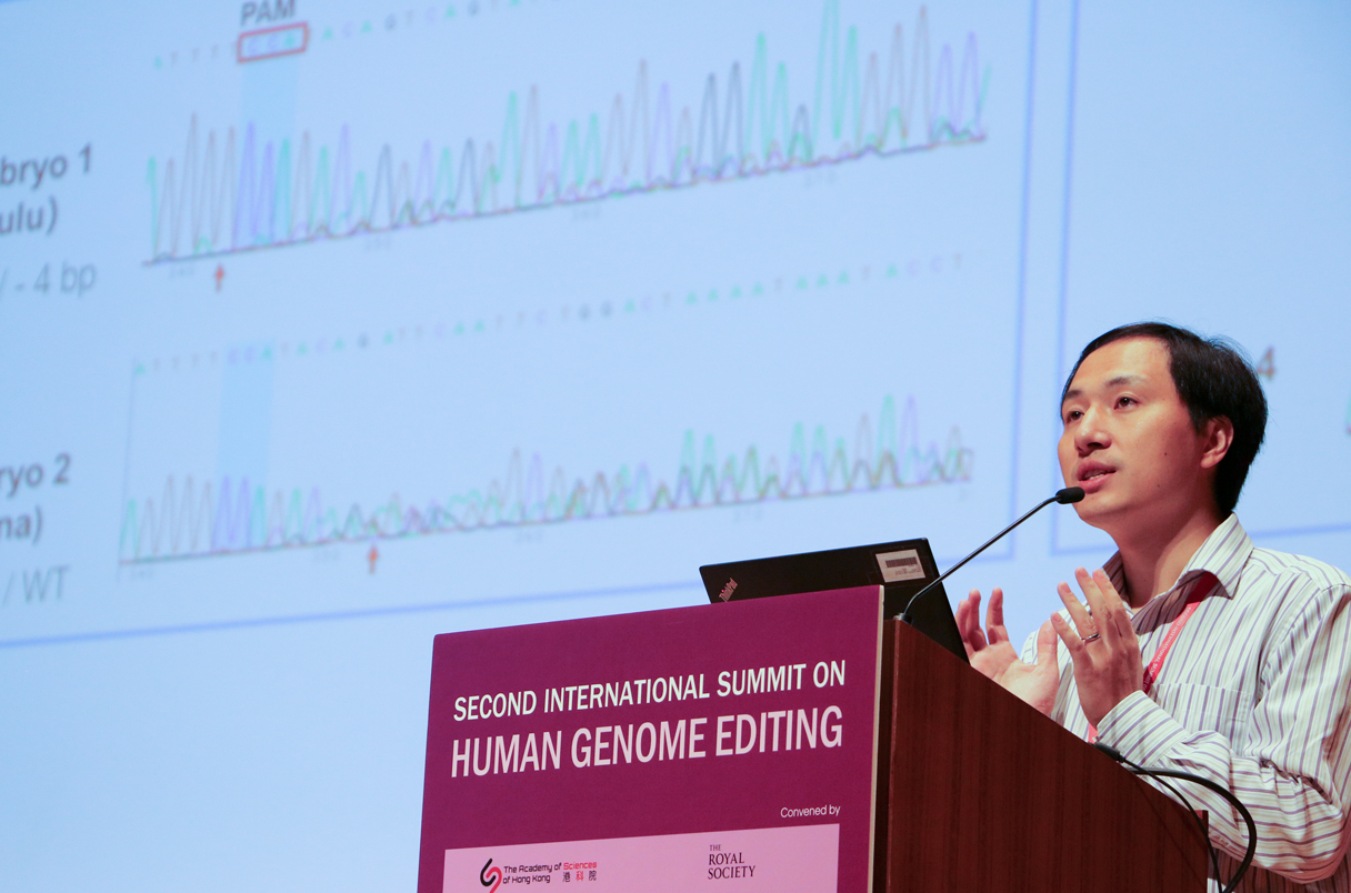 A person speaks at a podium during the Second International Summit on Human Genome Editing. A projected screen displays genetic sequences and data charts in the background.