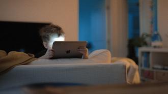 A child sitting on a couch in a dimly lit room, holding and looking intently at a tablet, as if testing the Goldilocks hypothesis for the perfect screen brightness.