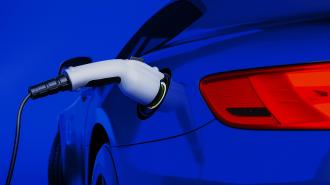 Close-up of an electric car being charged with a plug connected to its charging port at one of the many available charging stations. The blue car blends seamlessly into the blue background, easing any range anxiety drivers might have.