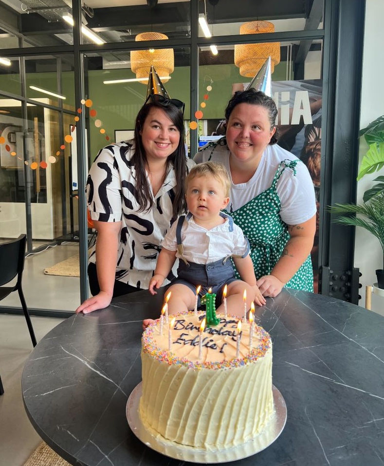 Two adults wearing party hats stand behind a seated child in front of a birthday cake with lit candles on a table. The cake features the text "Happy Birthday" and a number 1 candle.