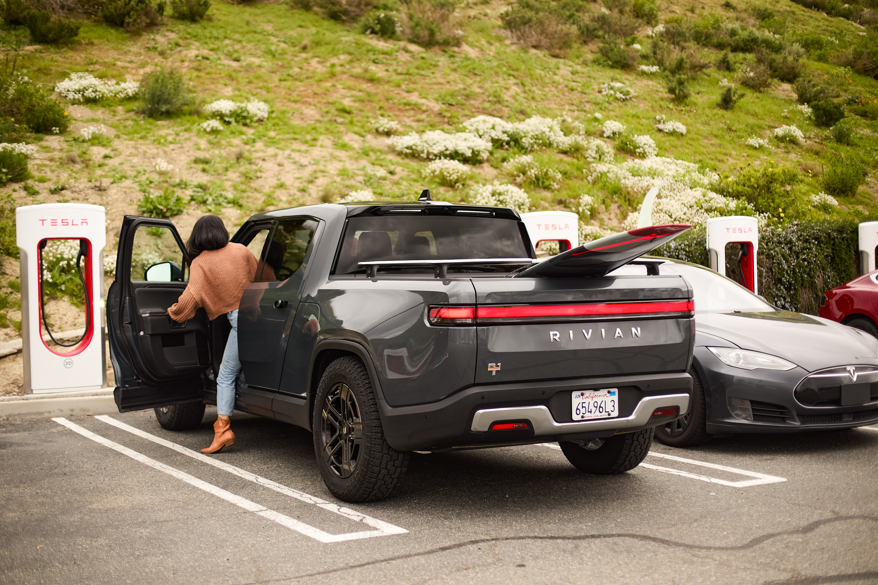 A person is getting into a Rivian truck parked at a Tesla charging station next to a Tesla car in a parking lot with greenery in the background.