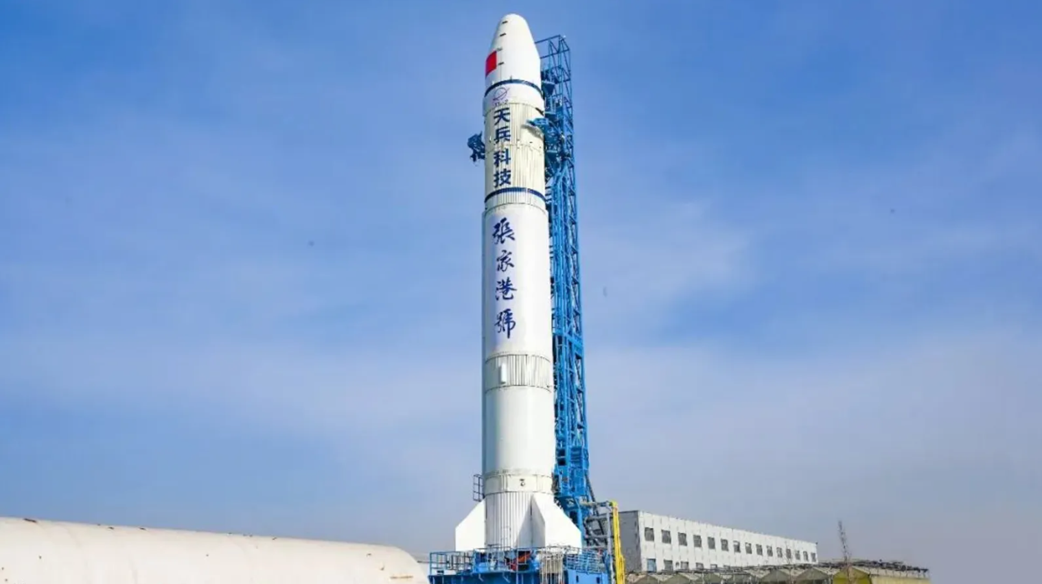 A white rocket stands vertically on a blue launch platform against a clear sky. Chinese characters are visible on the rocket's side.