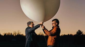 Two people hold a large white balloon outdoors at dusk, ready to release it and make sunsets an unforgettable memory. Trees and a fading sunset form the picturesque background.