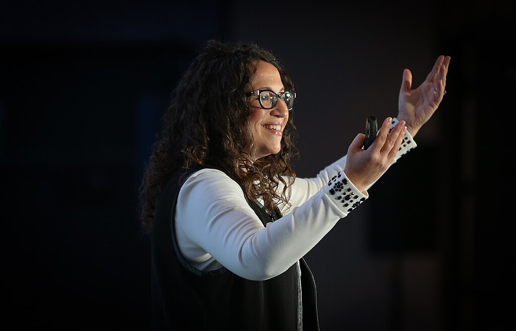 A woman with glasses gestures while speaking, holding a microphone, in a dimly lit room.