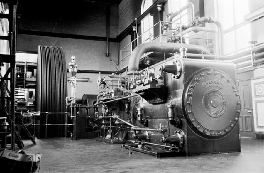 A large industrial steam engine with intricate pipes and gears inside a spacious room with tall windows.