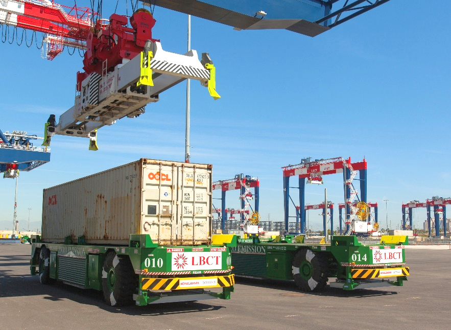 Automated vehicles at a port transport shipping containers beneath a crane on a sunny day.