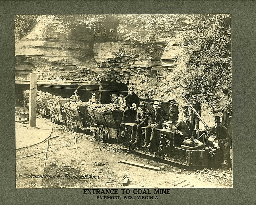 Historic photo of coal mine entrance in Fairmont, West Virginia. Miners sit on coal carts with a rocky tunnel in the background.