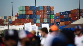 Stacks of colorful shipping containers at a port create a striking backdrop, while blurred people holding signs in the foreground hint at rising tensions over port automation amidst an ILA strike.