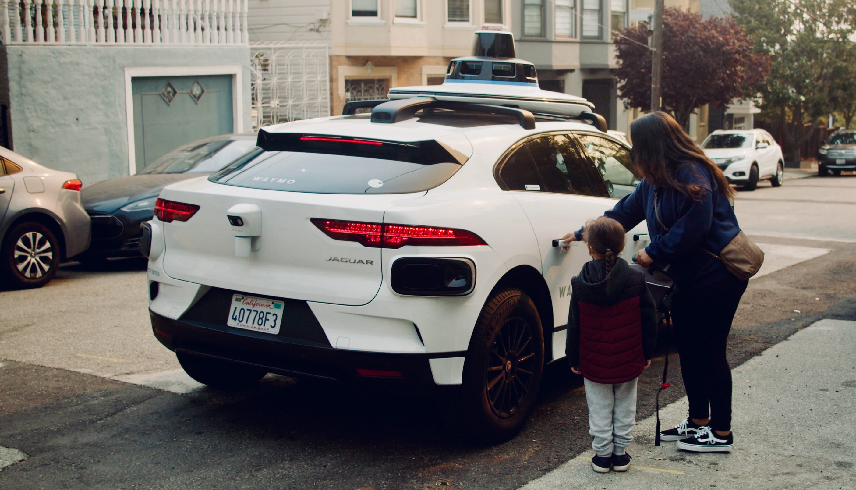 A woman and a child approach a parked white autonomous vehicle on a city street.