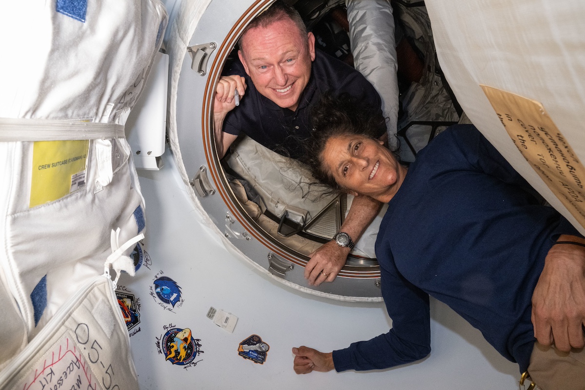 Two astronauts are floating in a spacecraft, smiling towards the camera through a circular doorway, with mission patches on the wall beside them.