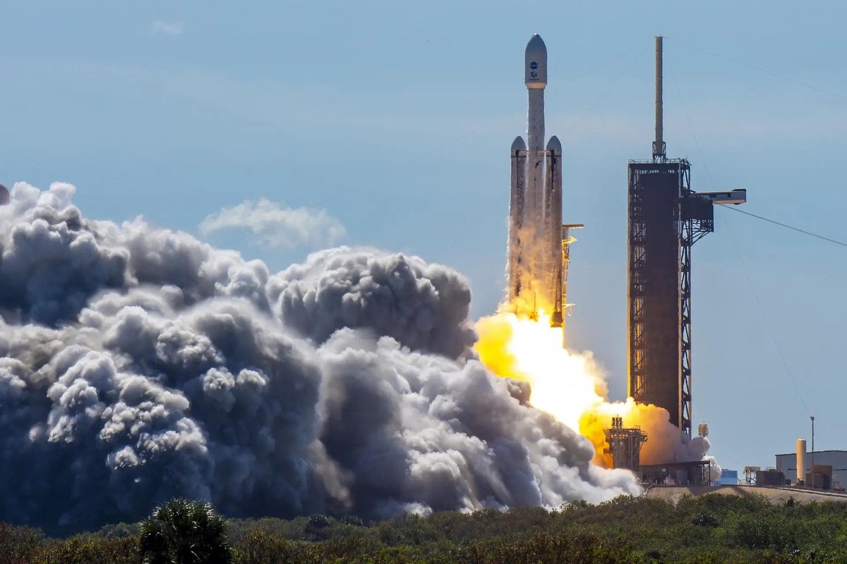 A rocket launches from a pad, surrounded by clouds of smoke and fire against a blue sky.