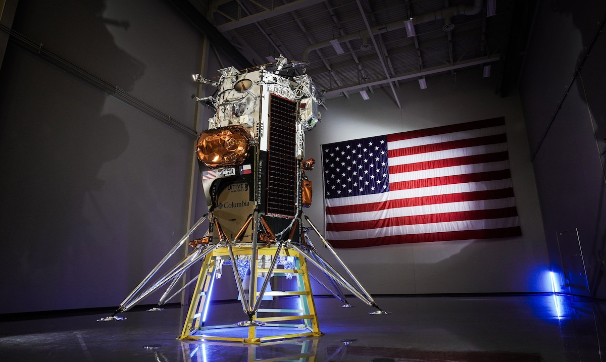 Spacecraft displayed indoors with an American flag in the background.