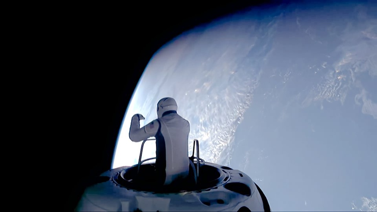 An astronaut in a white suit stands at the top hatch of a spacecraft as it floats near the edge of Earth, with the curve of the planet visible against the blackness of space.