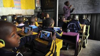 Children in a classroom use tablets and wear headphones while a teacher writes on a chalkboard.