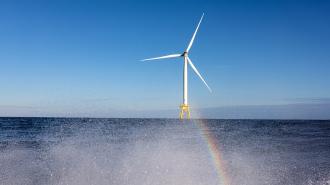 A wind turbine stands in the ocean under a clear blue sky, with a faint rainbow visible near the water's surface.