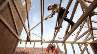 A construction worker balances on the wooden beams of a house frame, using a tool, with a red-tiled roof in the background under a blue sky, reflecting efforts to address the housing crisis.