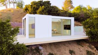 Modern white cube-shaped house with large glass windows, situated on a wooden deck on a hillside surrounded by trees and shrubs.